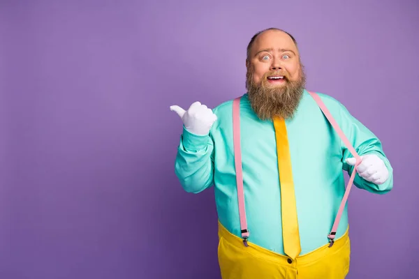 Portrait excited amazed fat bald man pull pink suspenders point forefinger copyspace recommend suggest select promotion ads wear teal yellow trousers isolated shine violet color background