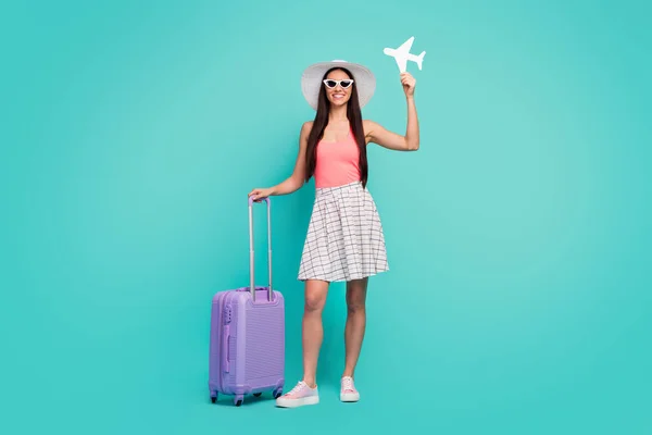 Full length photo of cheerful girl tourist hold white paper card plane enjoy summer weekend destination have suitcase wear sunglass sunhat sneakers isolated pastel teal color background — 图库照片
