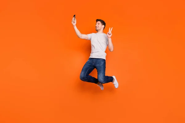 Pełna długość profil zdjęcie funky facet skakać wysoko w górę radując się zrobić selfie show v-znak symbol nosić paski t-shirt dżinsy trampki izolowane jasny pomarańczowy kolor tła — Zdjęcie stockowe