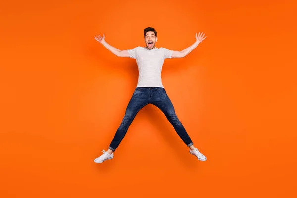 Full längd foto av galen funky kille hoppa högt upp särade händer ben gott humör öppen mun slitage randig t-shirt jeans sneakers isolerade ljusa orange färg bakgrund — Stockfoto