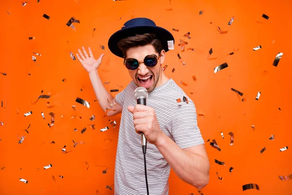 Foto di attraente pazzo funky ragazzo giovane canto karaoke utilizzando microfono coriandoli cadere indossare retro cappello sole specs strisce t-shirt isolato luminoso colore arancione sfondo — Foto Stock