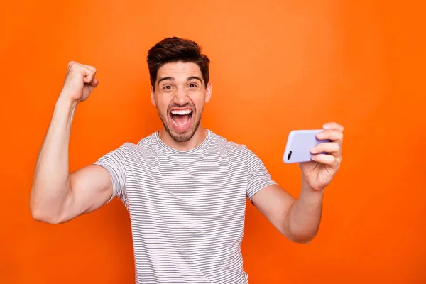 Zdjęcie fajnego podekscytowany facet dobry nastrój trzymać telefon podnieść pięść świętować pierwszy startup udany dochód nosić paski t-shirt odizolowany jasny pomarańczowy kolor tła — Zdjęcie stockowe