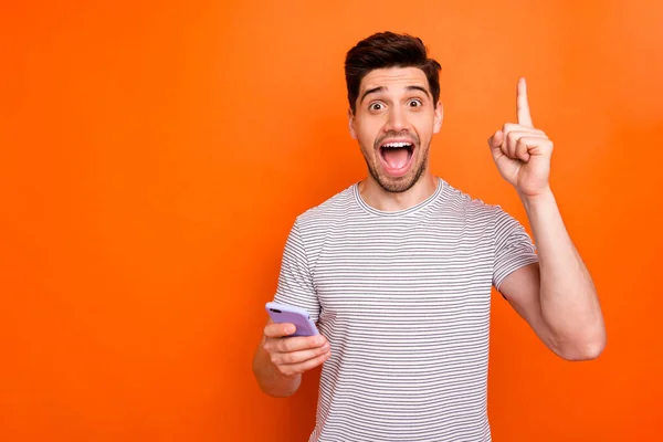 Photo of crazy millennial guy hold telephone hand raise index finger up have amazing creative blog post idea wear striped t-shirt isolated bright orange color background — Stockfoto