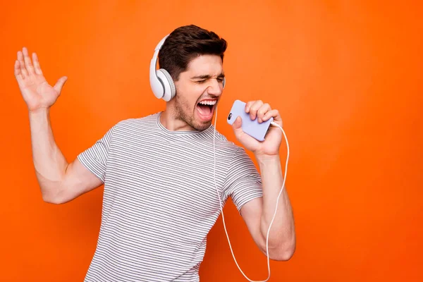 Photo of funky cool guy cheerful party mood chilling listening earphones hold telephone like microphone recording single wear striped t-shirt isolated bright orange color background — 스톡 사진