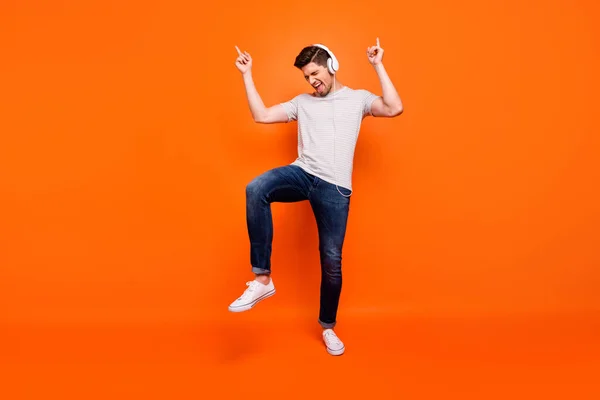 Full body photo of funky cool guy cheerful party mood chilling listening earphones direct fingers up empty space wear striped t-shirt jeans shoes isolated bright orange color background — Stockfoto