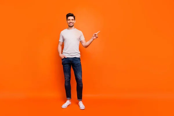 Full body photo of sales man guy cheerful person street casual look clothes directing finger empty space wear striped t-shirt jeans shoes isolated bright orange color background — Stockfoto