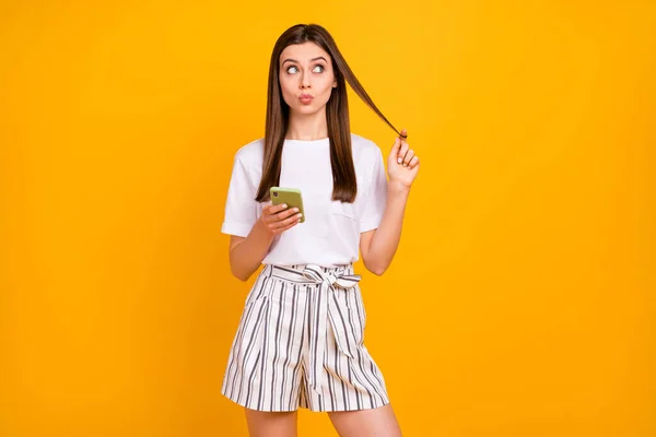 Photo of attractive pretty lady hold telephone look up empty space wounding curl around finger send shy kisses wear casual white t-shirt striped shorts isolated yellow color background — Stock Photo, Image