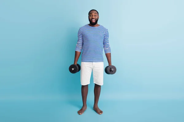 Full length photo of funny handsome dark skin guy holding two heavy dumbbells weight practicing hard outdoors wear striped sailor shirt shorts no shoes isolated blue color background — Stock fotografie