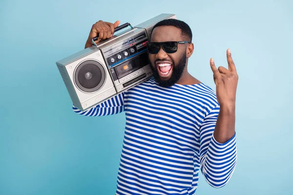 Hard rock. Photo of funny dark skin guy hold retro cassette recorder on shoulder showing metal horns hand wear sun glasses striped sailor shirt isolated blue color background — Stockfoto