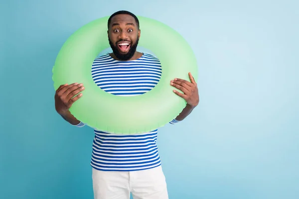 Photo of cheerful dark skin guy hold green life buoy around neck ready swim ocean sea traveler good mood sunny day wear striped sailor shirt isolated blue color background — Stock fotografie
