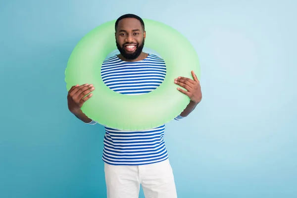 Foto de divertido alegre chico de piel oscura mantenga boya de vida verde alrededor del cuello listo nadar mar viajero buen humor día soleado usar camisa de marinero a rayas aislado color azul fondo — Foto de Stock