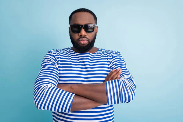 Photo of attractive dark skin guy arms crossed not smiling strictly looking travelers sea beach control wear sun glasses striped sailor shirt isolated blue color background — Stock Fotó