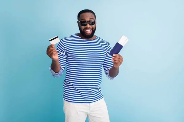 Foto de atractivo chico de piel oscura viajero comprar entradas con la ayuda de visa tarjeta de crédito usar gafas de sol rayas marinero camisa chaleco blanco pantalones cortos aislados de color azul de fondo — Foto de Stock