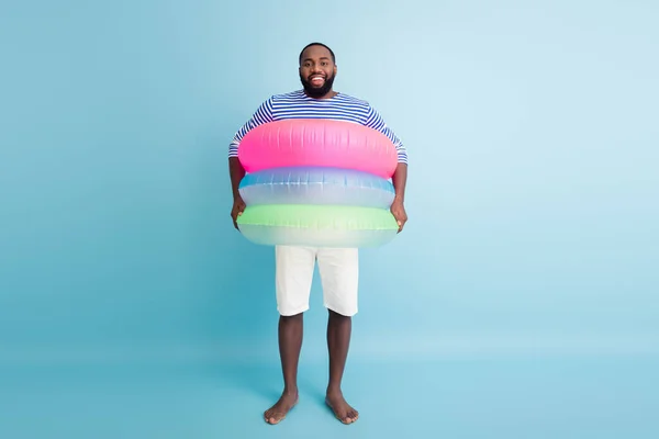 Full size photo of funky funny barefoot afro american guy enjoy weekend have colorful ring float lifesaver buoy ready swim in ocean wear white shorts frock isolated blue color background — Stockfoto