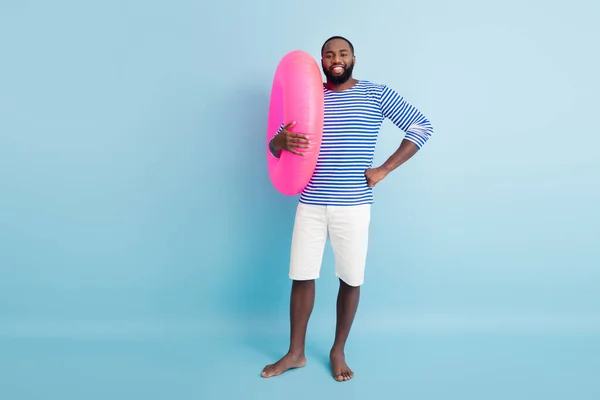 Full length photo positive brown hair barefoot afro american guy enjoy summer weekend hold pink float ring ready swim ocean wear nautical vest frock isolated blue color background — Stock fotografie