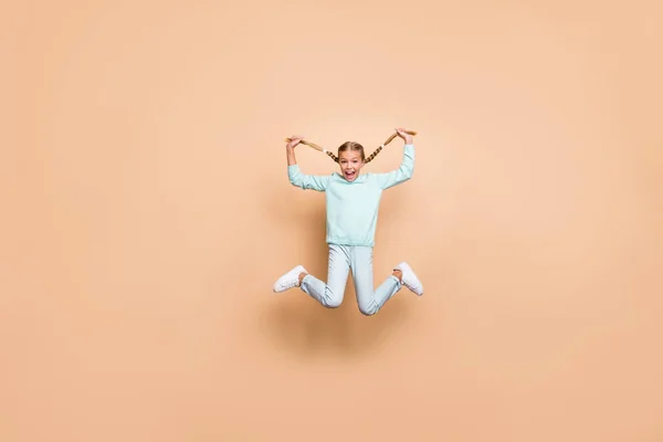 Full body photo of beautiful funny little lady jumping high up hold long braids hands excited good mood weekend wear blue sweater jeans footwear isolated beige color background — ストック写真