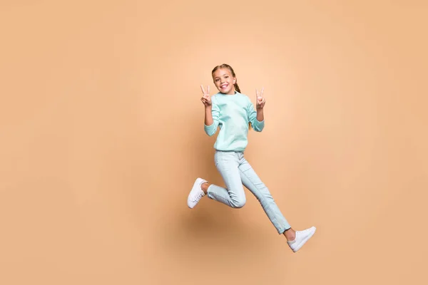 Full length photo of pretty beautiful funny little lady jumping high up showing v-sign symbols rejoicing weekend wear blue sweater jeans footwear isolated beige color background — Stok fotoğraf