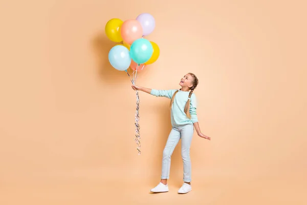 Full length profile photo of beautiful small lady hold many air balloons dreamy look up long braids wear blue sweater jeans sneakers isolated beige pastel color background — Φωτογραφία Αρχείου