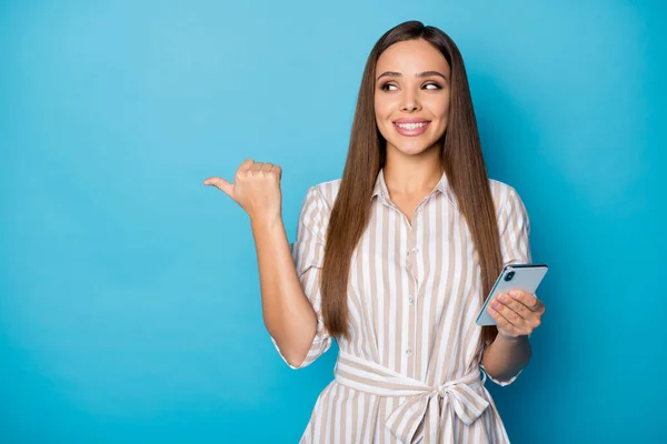 Venta. Foto de bastante atractiva blogger dama mantenga brazos telefónicos charlando amigos dirigiendo el dedo pulgar lado espacio vacío desgaste casual vestido a rayas aislado color azul fondo — Foto de Stock