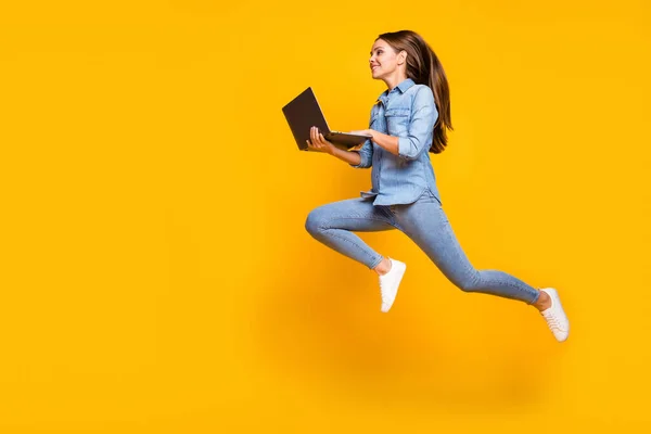 Full body profile photo of pretty business lady jump high holding notebook hands hurry work browsing laptop wear casual denim outfit white sneakers isolated yellow color background — 스톡 사진
