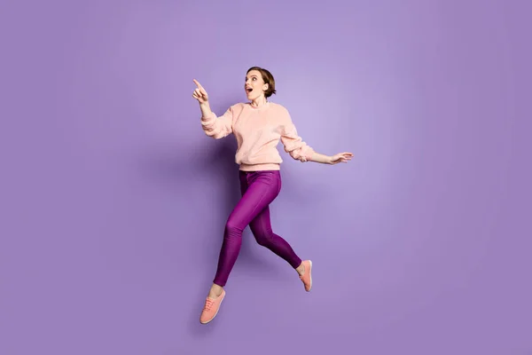 Full length photo of funny lady jumping up high see low shopping prices direct finger advert banner walking down street wear casual stylish pullover pants isolated purple color background — Zdjęcie stockowe
