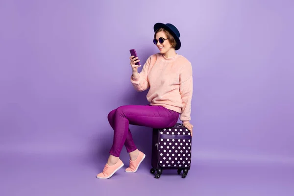 Πλήρες μέγεθος προφίλ φωτογραφία της συμπαθητικής κυρίας αναμονή πτήσης εγγραφής κρατήστε το τηλέφωνο κάθεται στην αίθουσα τσάντα φορούν καπέλο γυαλιά ηλίου casual μοντέρνο ροζ παντελόνι πουλόβερ απομονωμένο πορφυρό φόντο χρώμα — Φωτογραφία Αρχείου