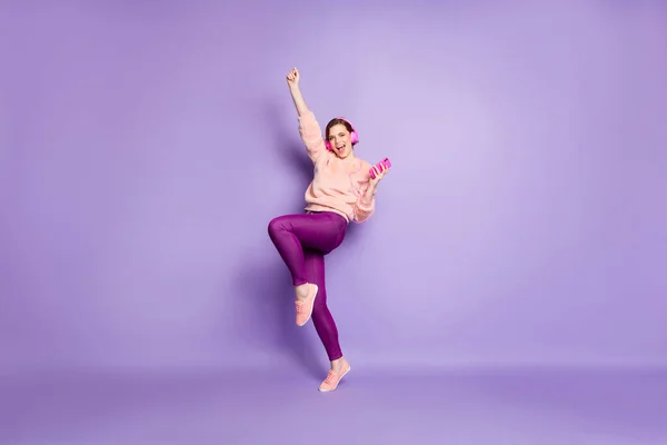 フルサイズの写真のファンキーな女性ホールド電話現代のイヤフォンで耳に耳を傾ける若者の音楽ダンス学生パーティー着用カジュアルなスタイリッシュなピンクプルオーバーパンツ孤立紫色の背景 — ストック写真