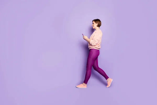 Full längd profil foto av vackra dam håller telefon chatta med vänner promenader ner gatan slitage casual elegant rosa pullover byxor isolerad lila färg bakgrund — Stockfoto