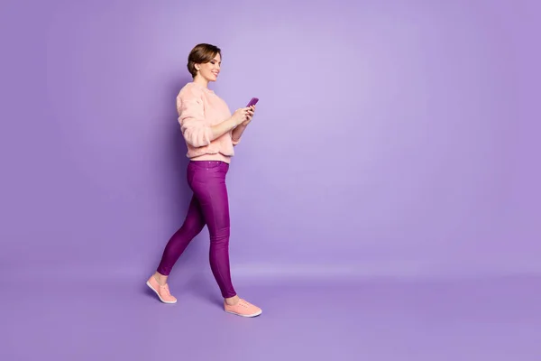 Jag är på väg. Full längd foto av vacker dam som håller telefon chatta med vänner promenader ner gatan slitage casual elegant rosa tröja byxor isolerad lila färg bakgrund — Stockfoto