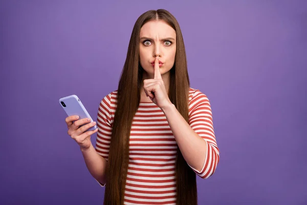 Тихіше не діліться. Серйозна молода дівчина використовує смартфон має приватне спілкування в соціальних мережах показують беззвучний знак індекс пальців губ носити білий смугастий джемпер ізольований фіолетовий кольоровий фон — стокове фото