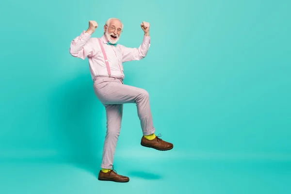 Full size photo of funny excited grandpa raise fists leg celebrate senior meeting party wear specs pink shirt suspenders bow tie pants shoes yellow socks isolated teal color background — 스톡 사진