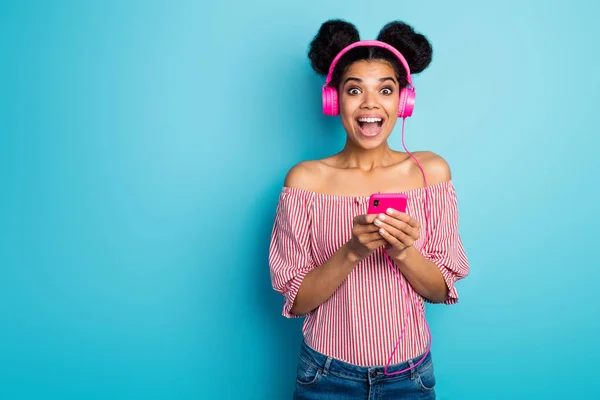 Photo of shocked dark skin lady open mouth hold telephone read good news listen music modern earphones wear red white striped shirt naked shoulders isolated blue color background — 스톡 사진