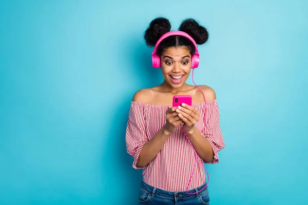 Photo of pretty shocked dark skin lady hold telephone read good news listen music modern technology earphones wear red white striped shirt naked shoulders isolated blue color background — 스톡 사진