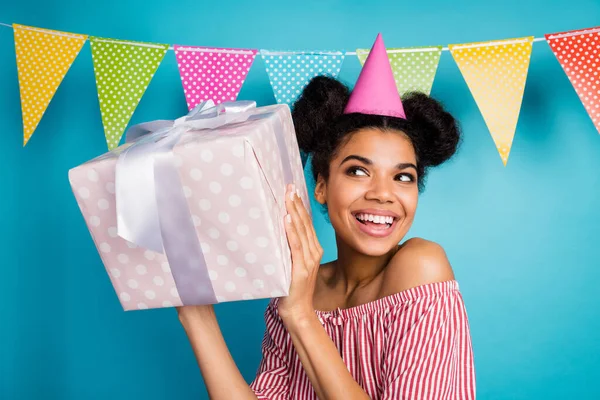Photo of funny curious dark skin lady hold large giftbox birthday girl party wear cone cap red white striped shirt naked shoulders colorful dotted flags hang over blue background — Zdjęcie stockowe
