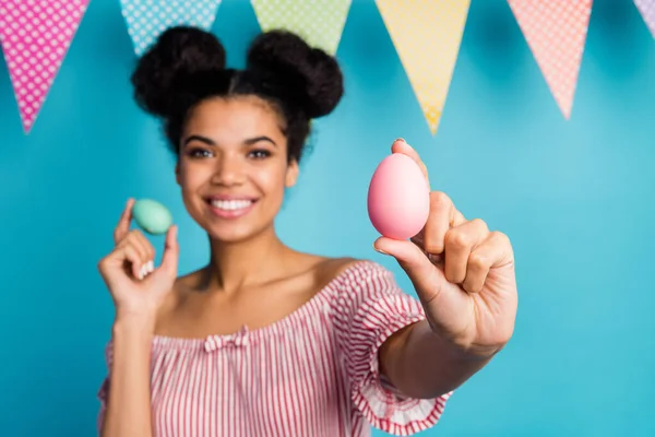 Photo of pretty dark skin lady showing holiday painted eggs colorful decor flags advice choose pink egg wear red white striped shirt naked shoulders isolated blue color blurry background