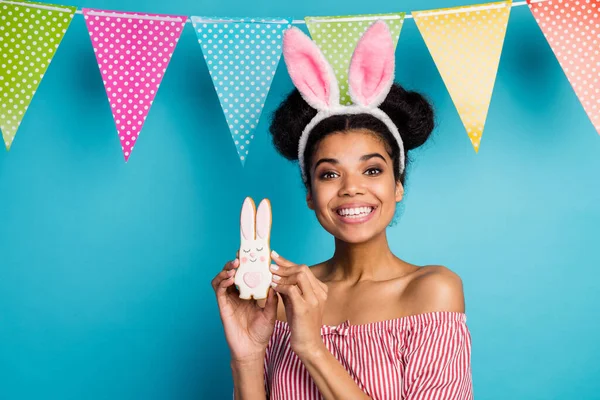 Photo of pretty dark skin lady holiday painted cookie bunny family dinner wear fluffy rabbit ears red white striped shirt off-shoulders colorful flags hang over blue color background — Zdjęcie stockowe