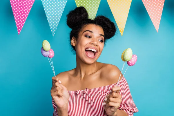 Photo of pretty dark skin lady hold holiday painted eggs on stick colorful decor flags good mood family meeting wear red white striped shirt naked shoulders isolated blue color background — ストック写真