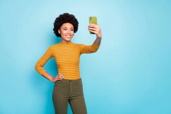 Foto de bela pele escura ondulada senhora segurar as mãos telefone fazendo selfies para novo instagram post bom humor desgaste amarelo listrado calças camisa isolado azul cor fundo — Fotografia de Stock