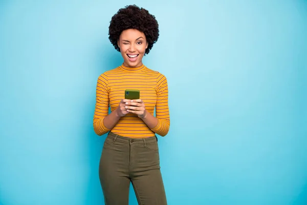 Foto de pele engraçada muito escura ondulado senhora segurar as mãos do telefone influenciador popular piscar os olhos brincalhão bom humor usar amarelo listrado calças camisa isolado azul cor fundo — Fotografia de Stock