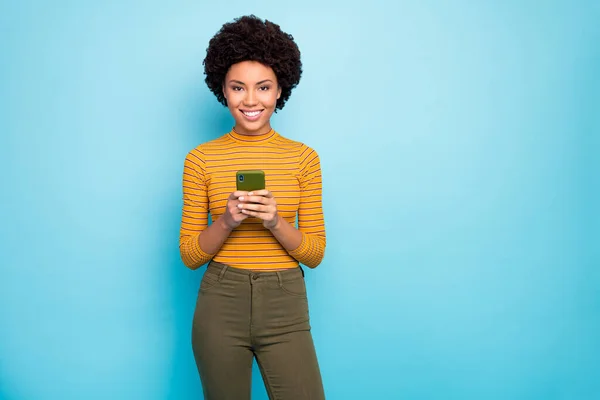 Photo de drôles de jolie peau sombre ondulée dame tenir les mains du téléphone lire les nouveaux commentaires influenceur populaire porter pantalon pull rayé jaune isolé fond de couleur bleue — Photo