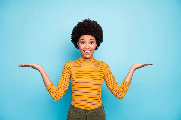 Foto de hermosa piel oscura dama ondulada mantenga las palmas abiertas presentando productos novedosos asesorando a precios bajos variantes usan pantalones de camisa de rayas amarillas aislados de color azul de fondo —  Fotos de Stock