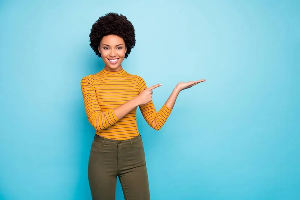 Foto de bela pele escura encaracolado modelo senhora segurar aberto palma novidade produto recomendando baixo preço dirigindo dedo desgaste amarelo listrado camisa calças isolado azul cor fundo — Fotografia de Stock