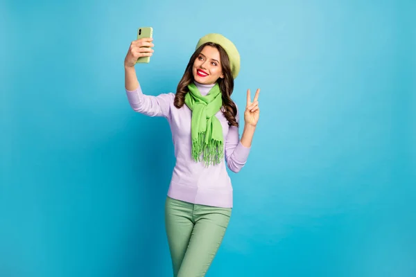 Foto di divertente affascinante signora tenere telefono facendo selfie che mostrano v-segno simbolo indossare alla moda verde berretto viola collo alto sciarpa pantaloni isolato colore blu sfondo — Foto Stock