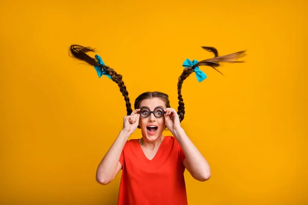 Foto van gekke grappige mooie dame student houd bril handen open mond goed gezichtsvermogen lange vlechten vlucht slijtage freak specs casual rood t-shirt geïsoleerd helder geel kleur achtergrond — Stockfoto