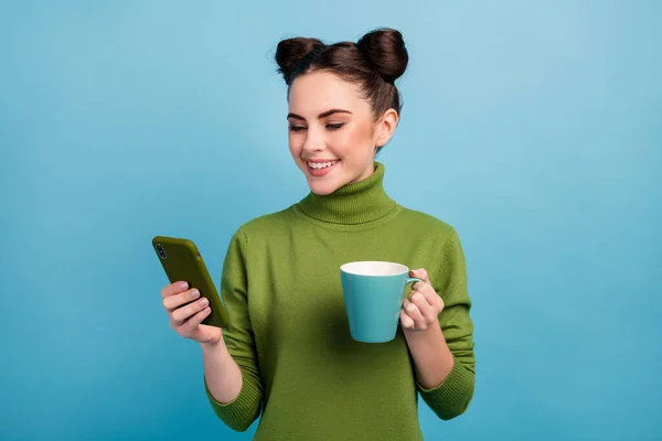 魅力的な十代の女性のホールド飲料カップの写真朝のフリーランスの仕事を閲覧電話ブログは暖かい緑のタートルネックプルオーバー隔離された青の色の背景を身に着けている — ストック写真