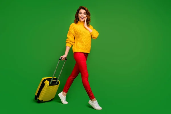 Полноразмерная фотография профиля взволнованной дамы, раскатывающей чемодан при регистрации на рейс авиакомпания меняет билеты на бизнес-класс в желтом свитере красные брюки туфли на зеленом фоне — стоковое фото