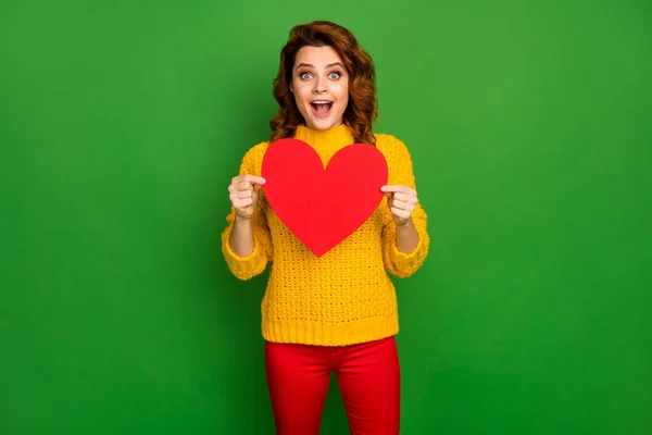 Foto de encantador ondulado animado senhora segurar papel coração forma cartão postal namorado convite para romance data desgaste amarelo malha camisola vermelho calças isolado brilhante verde cor fundo — Fotografia de Stock