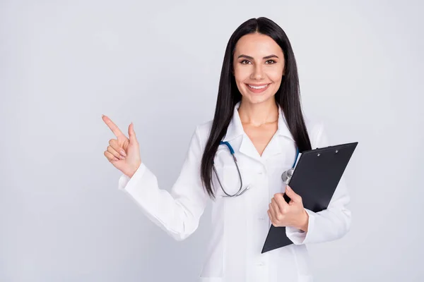 Foto de belo alegre doc senhora sorriso pacientes dedo direto vazio espaço seguro oferta segurar prancheta desgaste branco jaleco estetoscópio isolado cinza cor fundo — Fotografia de Stock