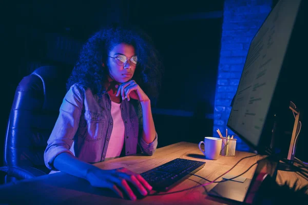 Profile photo of workaholic dark skin programmer lady look screen monitor check debugging system errors work overtime wear specs casual shirt sit table chair night office indoors