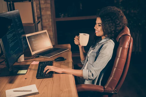 プロフィール側の写真正のプロの熟練したアフロアメリカの女の子のウェブ管理者は、職場のワークステーションで設計ホールドマグカップ飲料リラクゼーションを作成する夕方のテーブル作業バイナリコードに座る — ストック写真